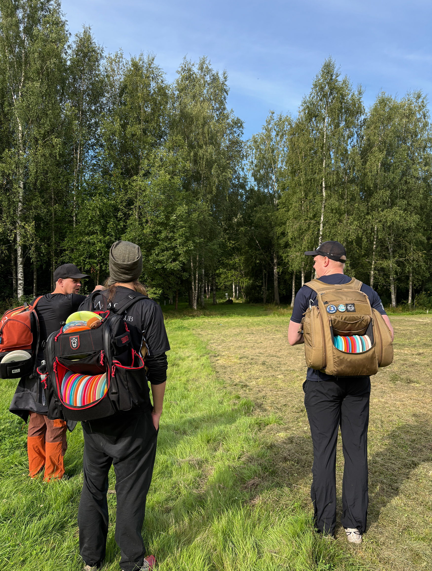 Kolme miestä seisovat nurmikolla selin kameraan ja keskustelevat. Taustalla koivumetsä.